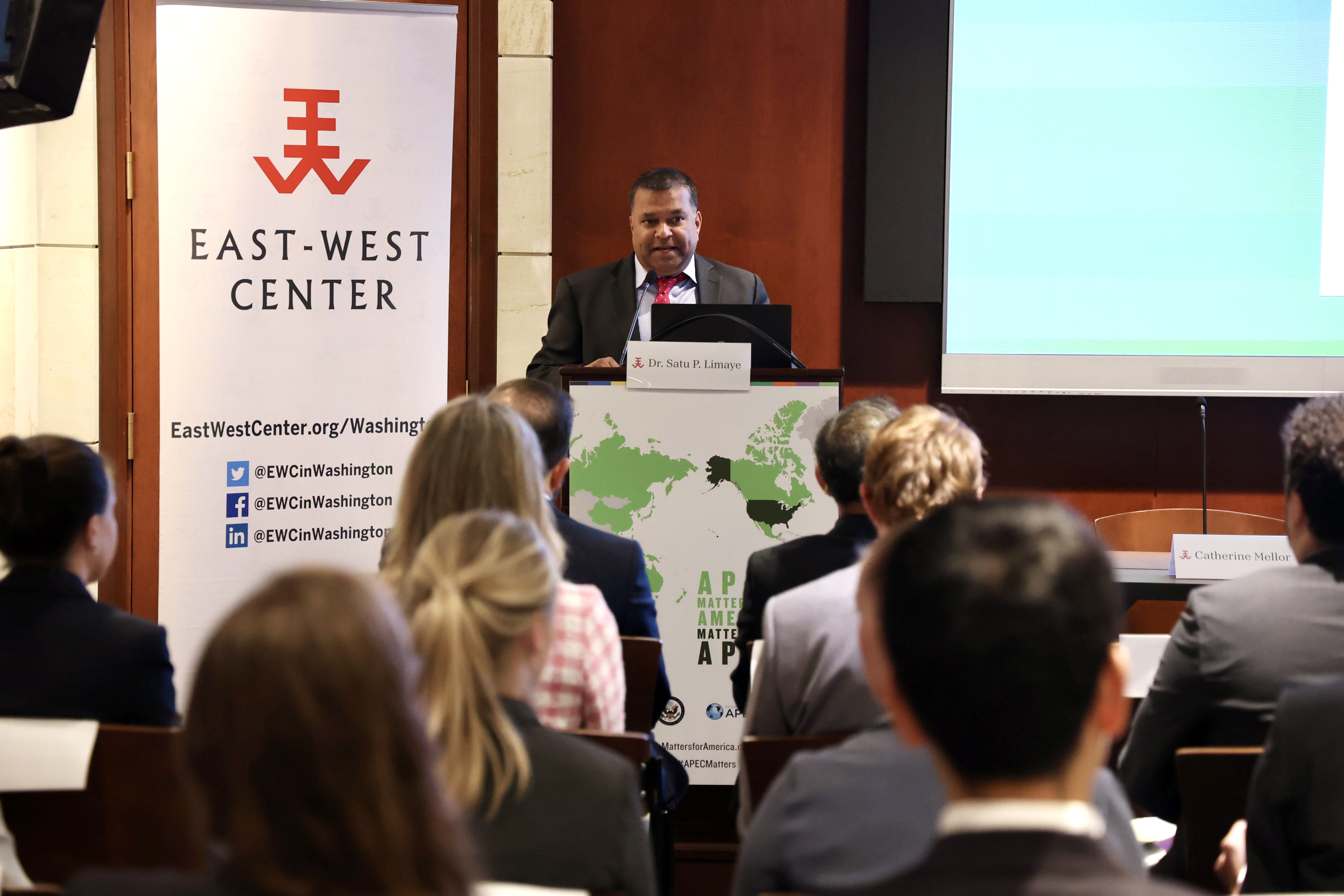 A man in a suit (Dr. Satu Limaye) speaks in front of an audience at a podium beside an East-West Center banner.