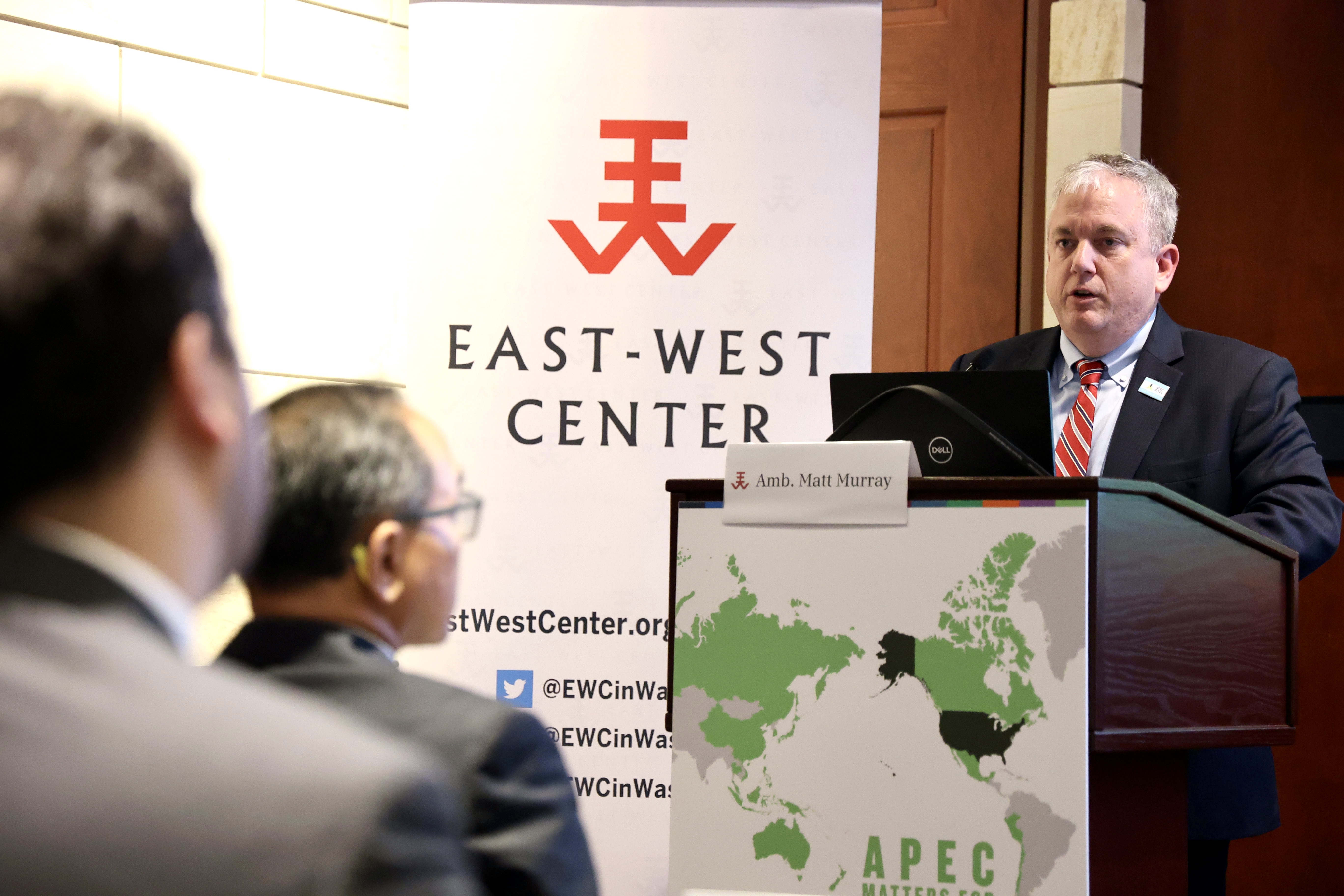 A man in a suit (Ambassador Matt Murray) speaks at a podium in front of an audience beside an East-West Center banner.