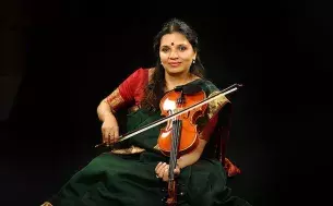 A smiling, dark skinned woman in colorful Indian clothing holding a violin on her left shoulder and a violin bow in her right hand