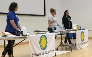 Three light-skinned women standing behind tables covered with disaster management supplies and Smithsonian Institute signage