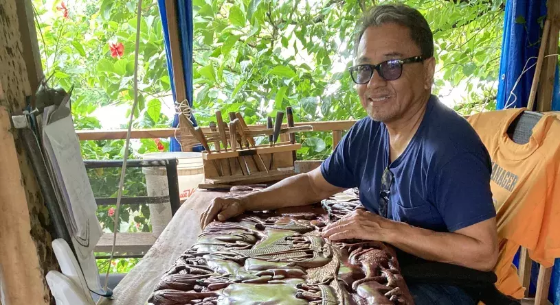 Smiling carver Dawrwin "Ling" Inabo, wearing a blue t-shirt and glasses, stands next to a large Palauan storyboard - a long wooden plank finely carved with images from a Palauan legend. Green foliage from trees shines brightly through the window in the background.