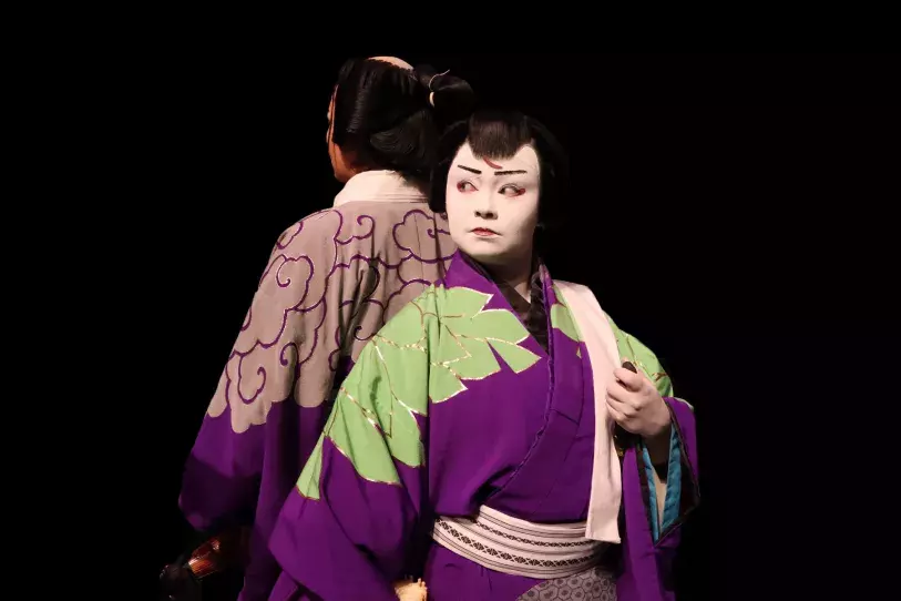 Photo of a kabuki actress wearing thick white facial makeup, black wig, and purple and green kimono robe, with another actor standing behind her and facing away, in similar dress and wig